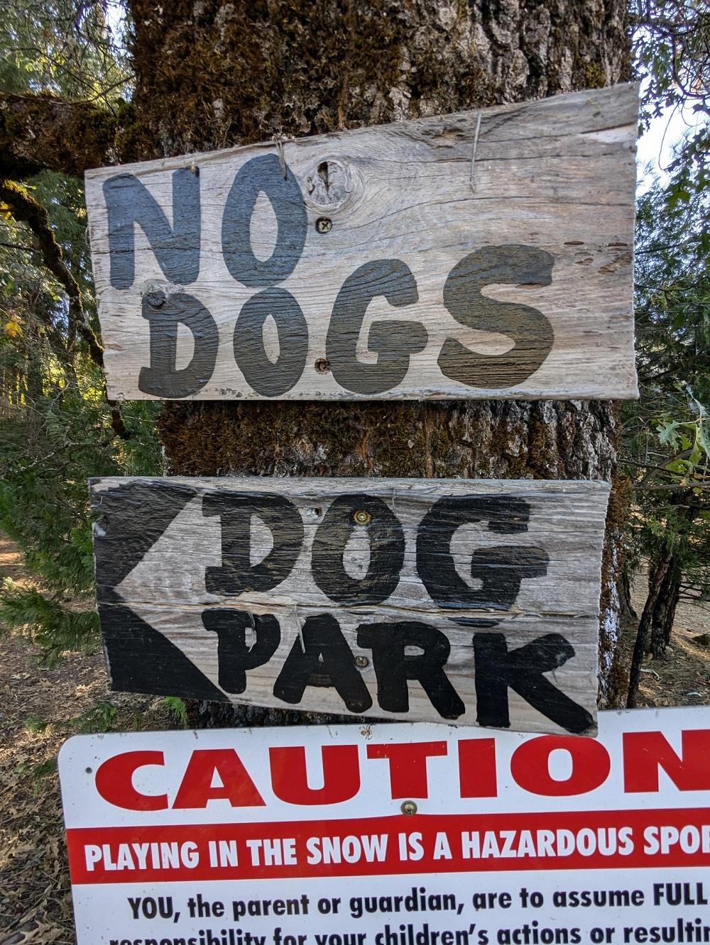 To vædede trætegn sømmet fast på et træ, hvoraf den ene står "NO DOGS" og den anden peger mod en "DOG PARK". Et advarselsskilt nedenfor advarer om at spille i sneen. Det er en visuel påmindelse om, at du ikke skal tage din hund med i parken.