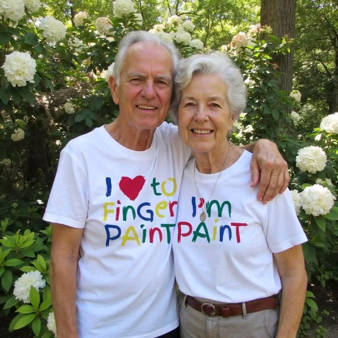 An elderly couple smiles, embracing, wearing matching white t-shirts that read "I ❤️ to Finger Paint, I'm Paint". The background features lush green foliage and white flowers.