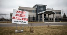 A sign reading "LAWSUIT ALLEGES SEXUAL ABUSE Spokane County Facilities" stands in front of the Spokane County Juvenile Detention Center, referencing the Spokane County lawsuit. The modern building is visible behind a fence on an overcast day.