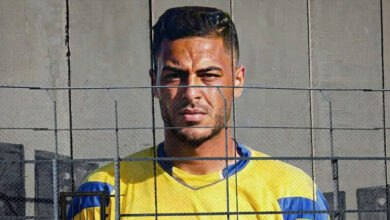 Saleem al-Ashqar, a Palestinian goalkeeper, stares intensely through a chain-link fence, wearing a yellow and blue jersey against a concrete wall. The image evokes themes of confinement and unfulfilled dreams.