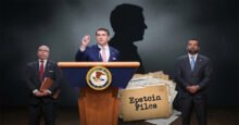 Todd Blanche speaks at a podium with the DOJ seal, flanked by two men, with a pile of "Epstein Files" and a shadowy silhouette behind him.