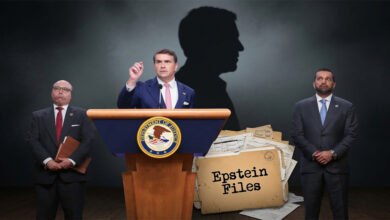 Todd Blanche speaks at a podium with the DOJ seal, flanked by two men, with a pile of "Epstein Files" and a shadowy silhouette behind him.