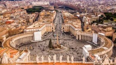 Aerial view of St. Peter's Square and the Vatican City, Rome, Italy. The large, oval square is filled with people, surrounded by colonnades and buildings. The central obelisk and surrounding architecture are key elements of the Vatican's history and connection to the Catholic Church.
