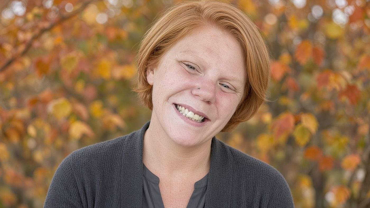 Danielle Grace Warren, a strawberry-blonde woman with a silver nose stud, smiles in a soft outdoor portrait.