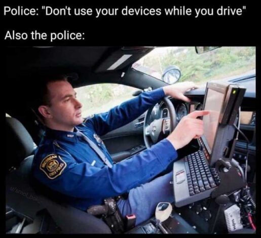 A Michigan State Police officer in uniform uses a touchscreen computer mounted inside a patrol car while driving, illustrating the hypocrisy of the "Don't use your devices while you drive" message.